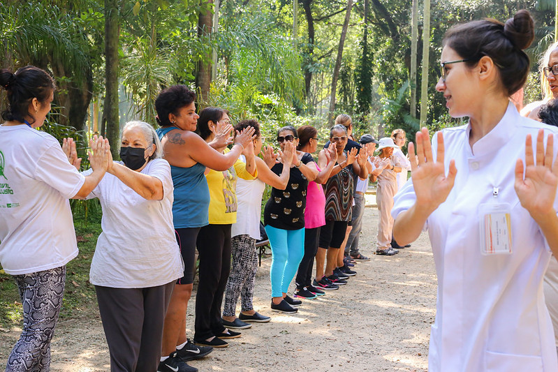 A imagem retrata uma atividade terapêutica ou de integração realizada ao ar livre, em um ambiente arborizado que se assemelha a um parque ou jardim público.Um grupo de pessoas, majoritariamente composto por mulheres adultas e idosas, está organizado em duas fileiras, uma de frente para a outra. Elas participam de uma dinâmica que envolve o contato físico ou o espelhamento de movimentos, com as palmas das mãos encostadas ou voltadas para as parceiras à frente. O grupo veste roupas leves e esportivas, adequadas para exercícios físicos. No lado esquerdo, uma das mulheres utiliza uma máscara facial preta.
