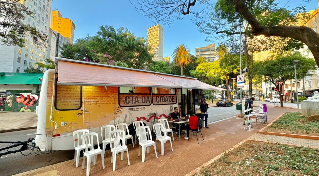 A imagem mostra uma unidade móvel de atendimento do CTA da Cidade (Centro de Testagem e Aconselhamento), estacionada ao lado de uma praça arborizada em uma área urbana. O veículo, do tipo trailer, ocupa o lado esquerdo da foto e possui uma grande cobertura retrátil aberta, criando uma área de espera na calçada. Em frente ao trailer há uma fileira de cadeiras plásticas brancas vazias. Próximo à entrada, duas pessoas estão sentadas em uma mesa, aparentemente realizando um atendimento ou cadastro, enquanto um profissional permanece em pé ao lado. Outra pessoa aparece mais à direita, próxima a um expositor com materiais informativos. Ao fundo, a paisagem é composta por árvores de grande porte, incluindo palmeiras, e edifícios altos, característicos da região central de uma grande cidade. A rua ao lado do trailer tem pouco movimento de veículos e pedestres. O ambiente transmite a ideia de uma ação de saúde pública realizada em espaço aberto, com estrutura organizada para acolhimento e atendimento da população.