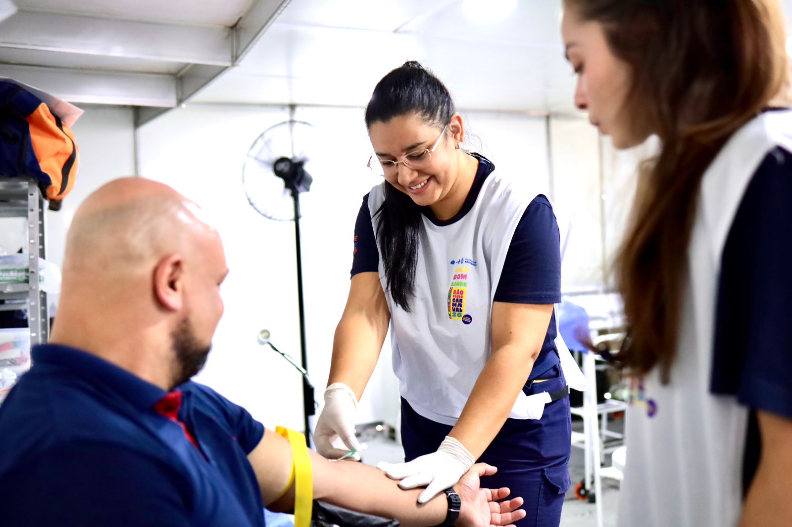 A imagem mostra um atendimento de saúde em um posto de saúde móvel, provavelmente uma tenda. No centro, uma profissional de saúde está sorrindo enquanto realiza um procedimento no braço de um homem sentado à esquerda. Ela usa luvas descartáveis e segura o braço dele com cuidado, sugerindo que está coletando sangue ou aplicando algum tipo de injeção. O homem, que é careca e tem barba, veste uma camiseta azul e mantém o braço estendido, com uma faixa amarela ajustada acima do cotovelo, típica de coleta de sangue. À direita, há outra mulher, também com uniforme semelhante, observando o procedimento, possivelmente auxiliando ou acompanhando o atendimento. O ambiente ao fundo é claro e organizado, com equipamentos médicos e uma luminária visível, reforçando a ideia de um espaço clínico temporário.