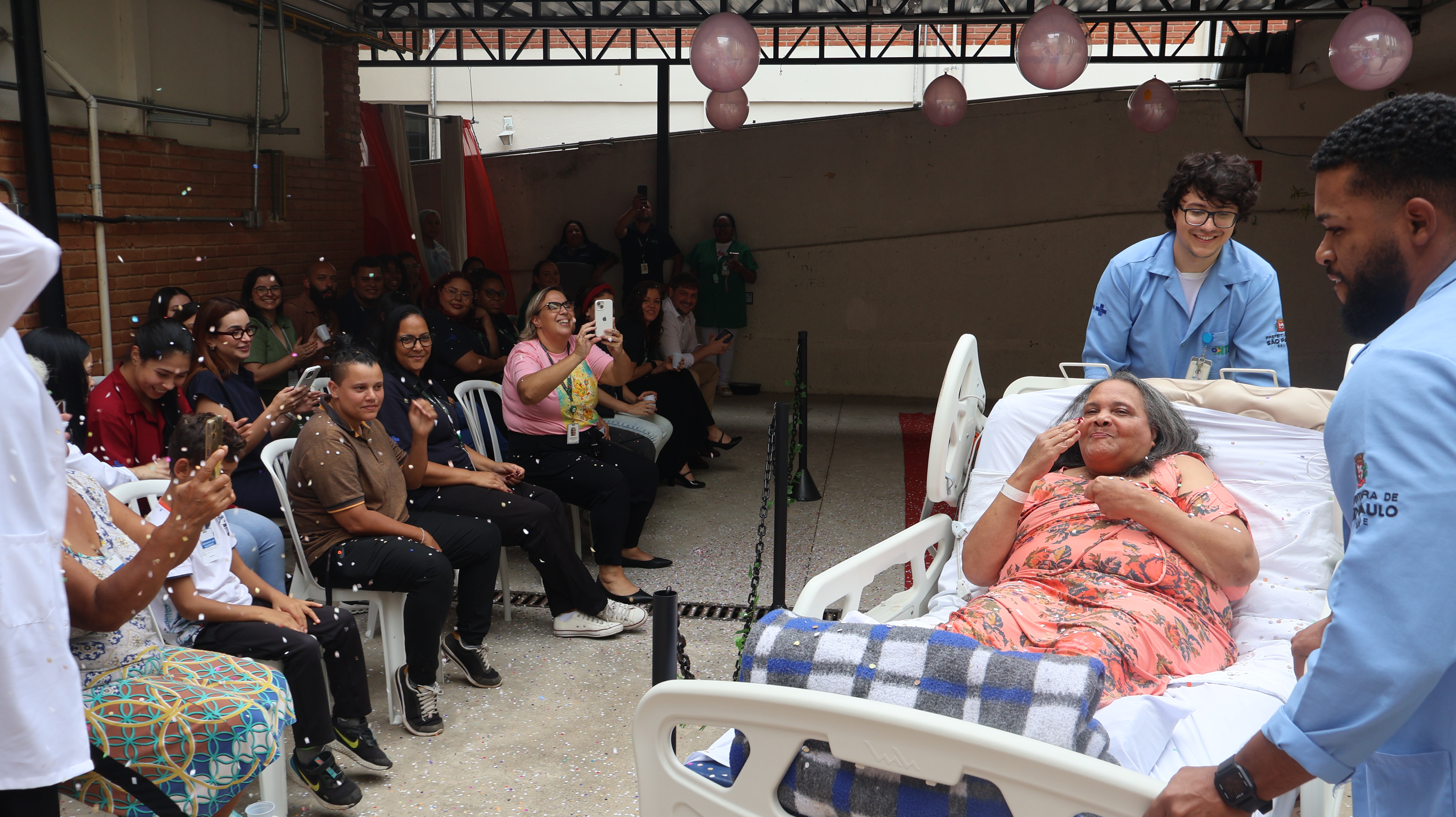 A fotografia registra um evento festivo em uma área externa ou pátio coberto do Hospital Municipal Guarapiranga. No centro da composição, uma paciente idosa está deitada em uma cama hospitalar branca, vestindo um robe floral rosa e coberta por uma manta xadrez azul. Ela sorri e faz um gesto de beijo para a plateia, visivelmente emocionada.  A cama é conduzida por dois profissionais de saúde vestindo jalecos azuis claros com o logotipo da Prefeitura de São Paulo. Ao lado esquerdo, uma plateia formada por funcionários e familiares está sentada em cadeiras brancas, aplaudindo, sorrindo e registrando o momento com celulares. O ambiente está decorado com balões cor-de-rosa no teto e confetes brancos caem sobre a paciente, criando uma atmosfera de desfile e celebração.