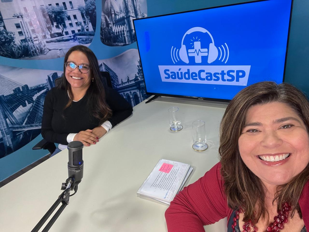 A imagem mostra duas mulheres sorrindo em um ambiente que parece ser um estúdio de gravação de podcast ou conteúdo em áudio. Ambas estão sentadas em torno de uma mesa, onde há um microfone de gravação, dois copos de água e anotações impressas.  Ao fundo, um monitor exibe a identidade visual “SaúdeCastSP”, acompanhada de um ícone de fones de ouvido com um símbolo de saúde ao centro, sugerindo que o espaço é voltado à produção de conteúdo sobre saúde. A parede também possui imagens em tons de azul que remetem à cidade de São Paulo e a cenários urbanos.  A cena transmite um clima de conversa, entrevista ou gravação de programa institucional, com um ambiente acolhedor e profissional voltado à comunicação em saúde.