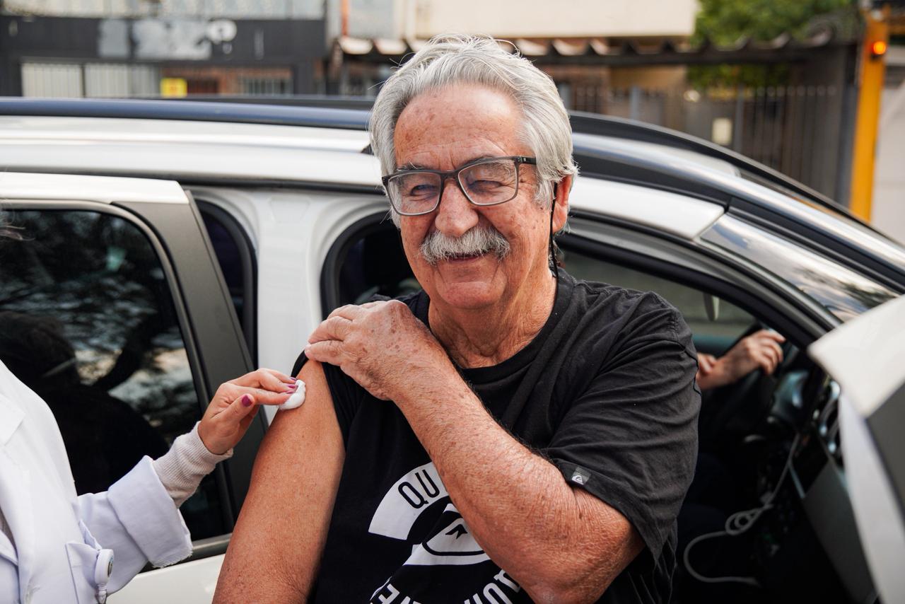 Homem idoso, de cabelos grisalhos e óculos, sorri enquanto recebe uma vacina no braço esquerdo, ao lado de um carro estacionado em sistema drive-thru. Ele veste camiseta preta e segura a manga da roupa para facilitar a aplicação. A profissional de saúde aparece parcialmente na imagem, aplicando o curativo no local da vacina. Ao fundo, há elementos urbanos desfocados.