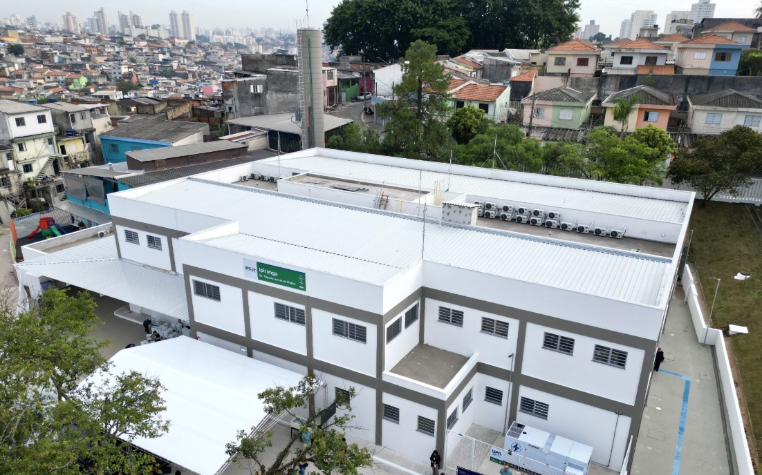 A imagem mostra uma unidade de saúde de médio porte, vista de cima, em um bairro urbano. O prédio é novo, com pintura branca e detalhes em cinza, telhado metálico e diversos aparelhos de ar-condicionado instalados na parte superior. Na fachada lateral, há uma placa verde com o nome UPA Ipiranga – Unidade de Pronto Atendimento 24h. Em frente ao prédio, foi montada uma estrutura com cobertura branca, provavelmente para abrigar visitantes ou a realização de um evento inaugural. O entorno é composto por casas simples e coloridas, dispostas em ruas estreitas e irregulares, com uma área de maior adensamento urbano ao fundo, onde se veem prédios residenciais mais altos. A cena sugere a integração da unidade de saúde com a comunidade local, em uma região de grande densidade populacional.