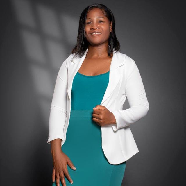 A imagem mostra uma mulher em pé, posando para um retrato em estúdio, com expressão serena e um leve sorriso. Ela veste um blazer branco sobre um vestido ou conjunto na cor verde-petróleo e segura a lapela do blazer com uma das mãos, enquanto a outra permanece relaxada ao lado do corpo. Seus cabelos são lisos, na altura dos ombros, repartidos de lado, e ela está voltada para a câmera. O fundo é cinza-escuro, com iluminação suave que cria sombras discretas, destacando a figura da retratada. A composição é simples e elegante, típica de um retrato profissional ou institucional, transmitindo uma imagem de confiança, acolhimento e profissionalismo.