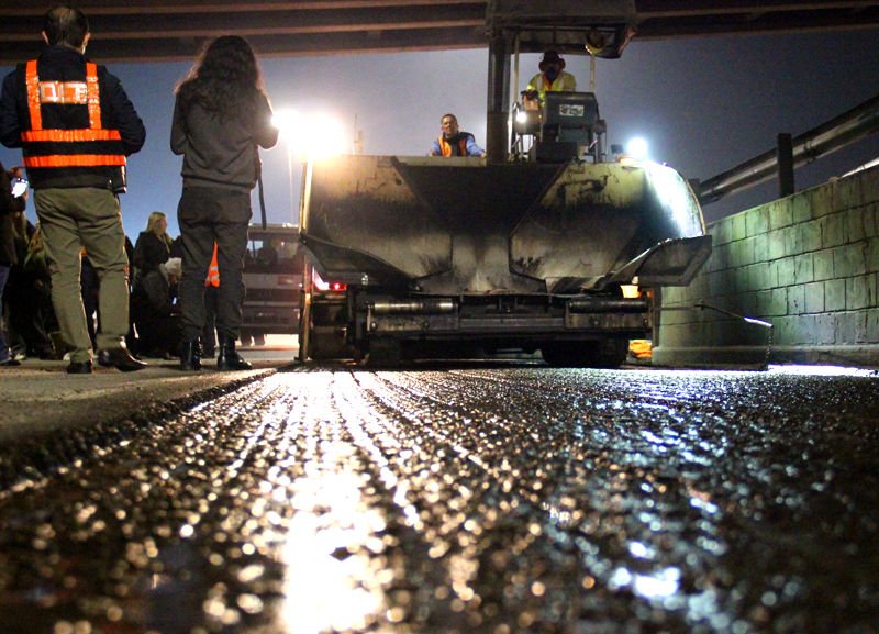 A imagem mostra uma cena noturna de obra viária vista bem de perto do nível do asfalto. O primeiro plano está em foco e revela o piso áspero e úmido, possivelmente recém-fresado ou preparado para recapeamento, com brilho de pequenas gotas refletindo a luz. Ao fundo, desfocado, há um caminhão branco com faróis acesos apontando para a câmera e, ao lado dele, cones de sinalização laranja e trabalhadores vestindo uniformes fluorescentes em tons de laranja e amarelo. A iluminação sugere que o serviço acontece à noite, com luzes artificiais e reflexos brilhando na pista.