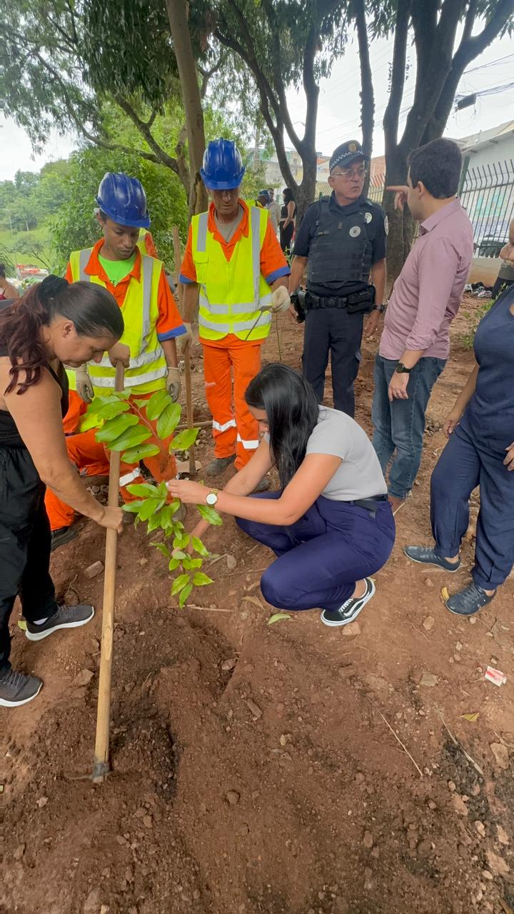 Duas Estudantes agachadas remexem na terra e uma delas usando enxadas realizam o plantio de muda. Ao lado dois zeladores usam uniformes na cor  laranja, capacetes na cor azul, observam o plantio e estão ladeados pelo policial da GCM e pelo subprefeito da Cidade Tiradentes.