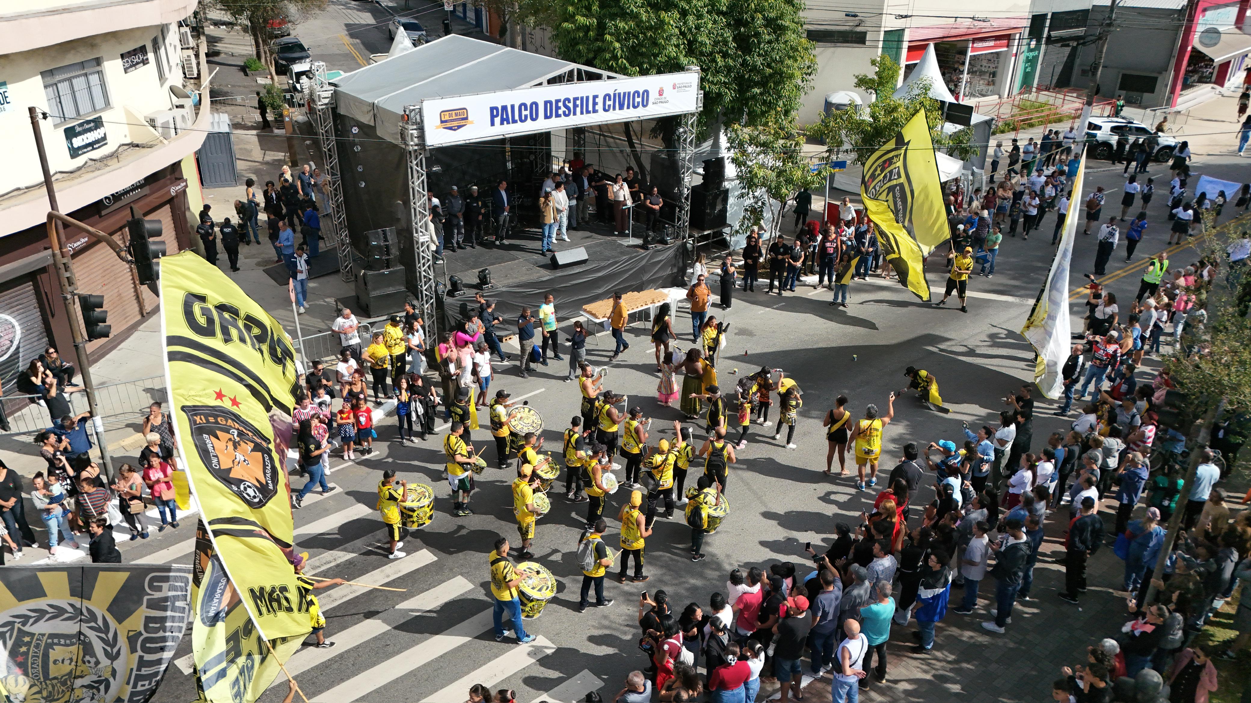 Uma imagem aérea de um desfile cívico em uma rua movimentada. Ao centro, um palco cinza com a inscrição 
