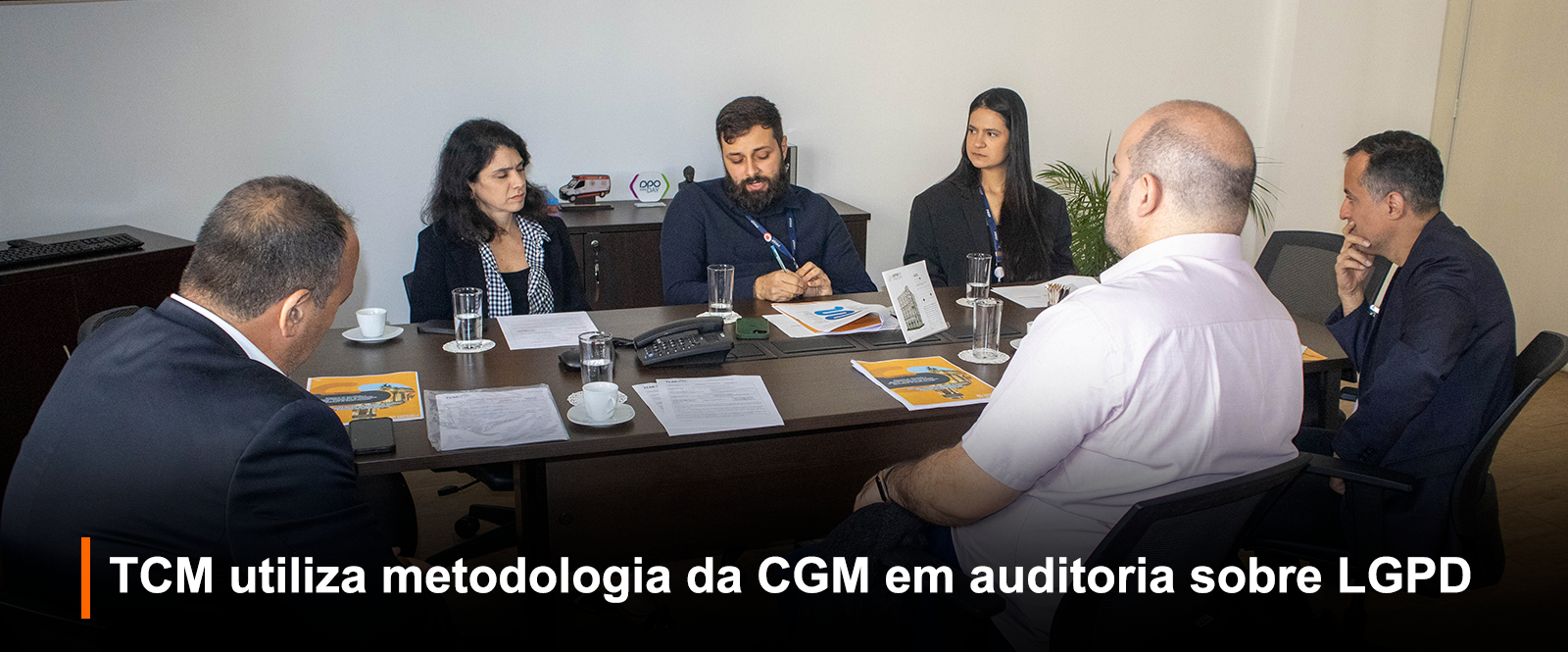Fotografia de reunião em ambiente fechado: seis pessoas ao redor de uma mesa de madeira. Um homem de barba escura e camisa preta gesticula enquanto fala, com papéis e revista à frente. Outros participantes escutam atentamente. Na mesa, copos, xícaras e documentos. Ao fundo, parede clara com logotipo 