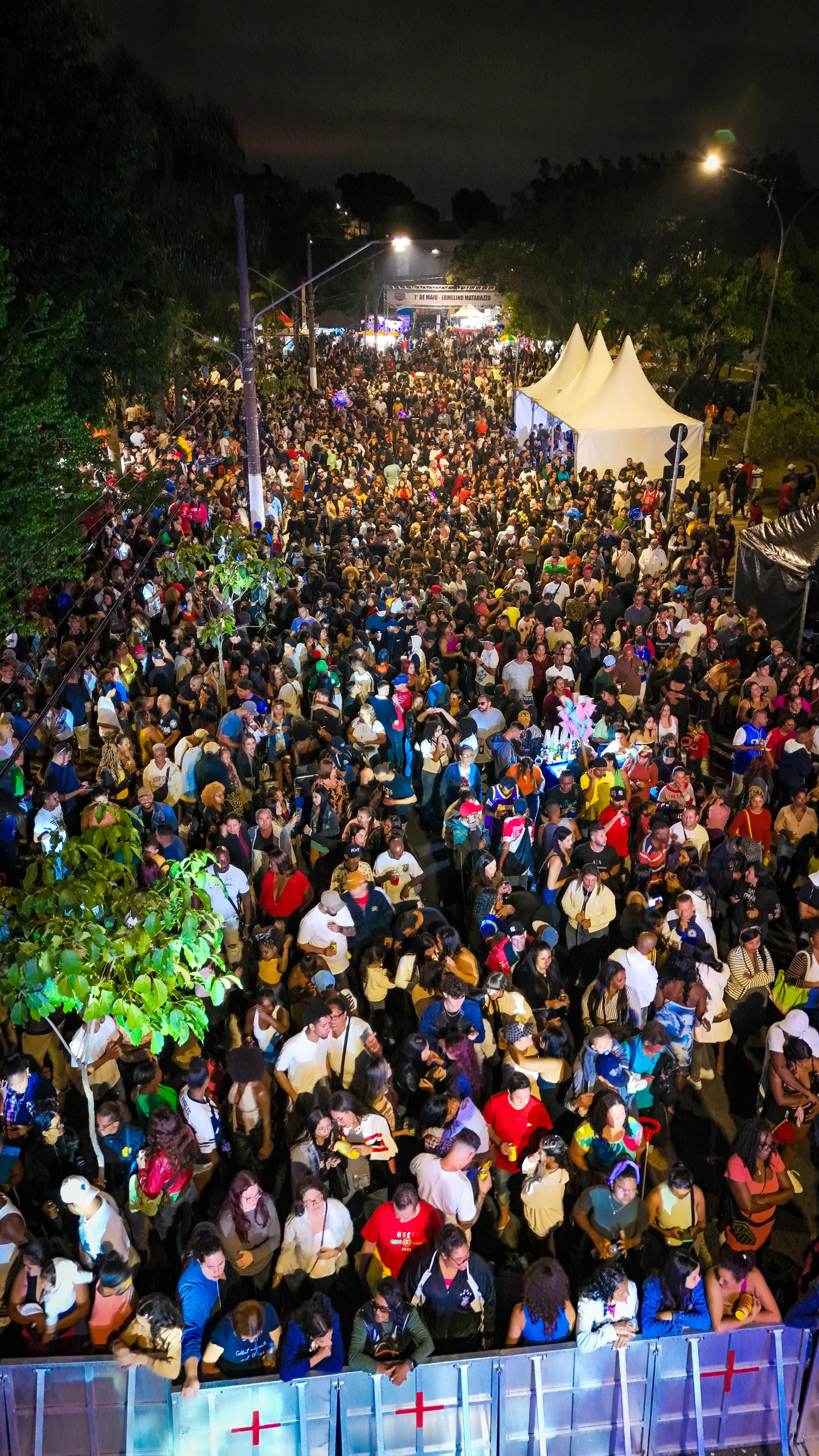 Uma vista aérea de uma multidão densa, ocupando uma rua e calçadas durante um evento noturno. A rua é ladeada por árvores com folhagem escura, e ao fundo, tendas brancas com telhados pontudos se destacam no meio da aglomeração. A iluminação ambiente é escassa, vindo principalmente de postes de luz e de algum brilho distante, realçando a grande quantidade de pessoas que parecem estar se divertindo em um festival ou celebração. No primeiro plano, parte de uma estrutura de metal, possivelmente uma barreira ou palco, é visível, com alguns pontos vermelhos em forma de "mais" desenhados nela.