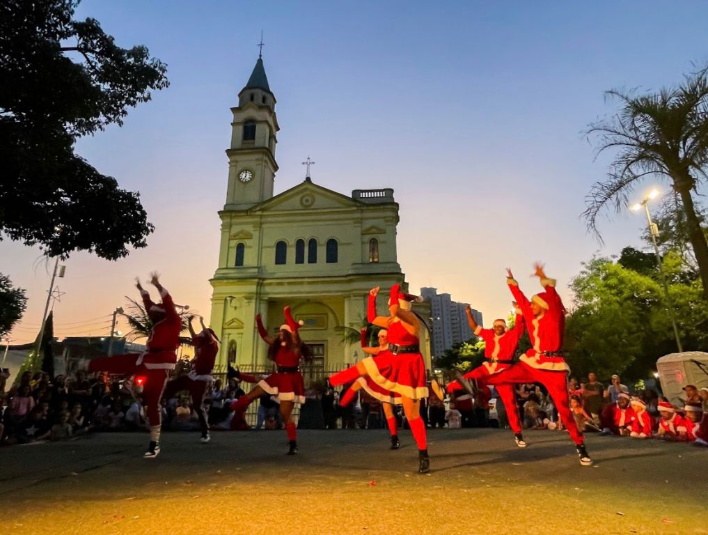 #Pracegover: foto ao ar livre capturada ao entardecer, exibe uma performance vibrante de um grupo de sete dançarinos vestidos com trajes natalinos (vermelho e branco, remetendo a ajudantes do Papai Noel), que estão em poses dinâmicas com os braços erguidos em frente a uma igreja de cor clara, que domina o fundo com sua torre sineira e relógio. O céu está em tons de crepúsculo azul e laranja, e no primeiro plano, uma multidão escura está sentada no chão, assistindo à apresentação, emoldurada por árvores escuras nas laterais.