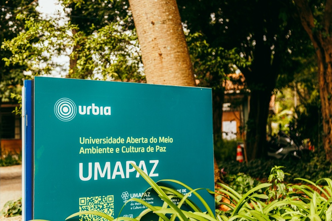 Placa institucional em tom verde-azulado com o nome UMAPAZ – Universidade Aberta do Meio Ambiente e Cultura de Paz escrito em destaque. No topo aparece a marca “urbia”. A placa está instalada em área externa, cercada por vegetação verde, com folhas e plantas em primeiro plano. Ao fundo, vê-se o tronco de uma árvore e um ambiente arborizado, indicando que o local fica em um parque ou espaço natural.