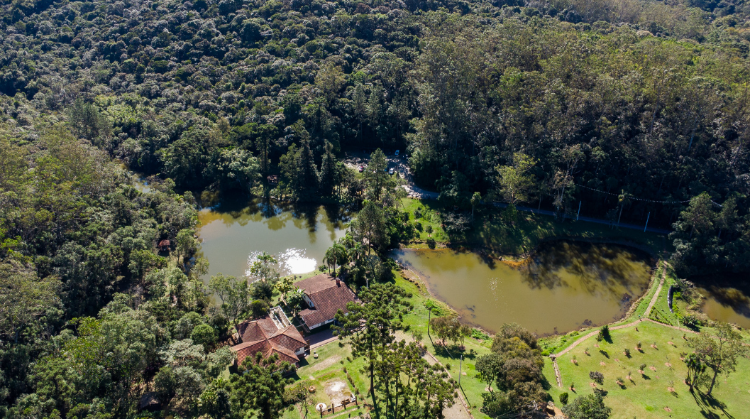 Vista aérea de um parque com densa área de mata ao redor, dois lagos de água esverdeada conectados e, ao lado, uma construção com telhado vermelho cercada por jardins e caminhos.