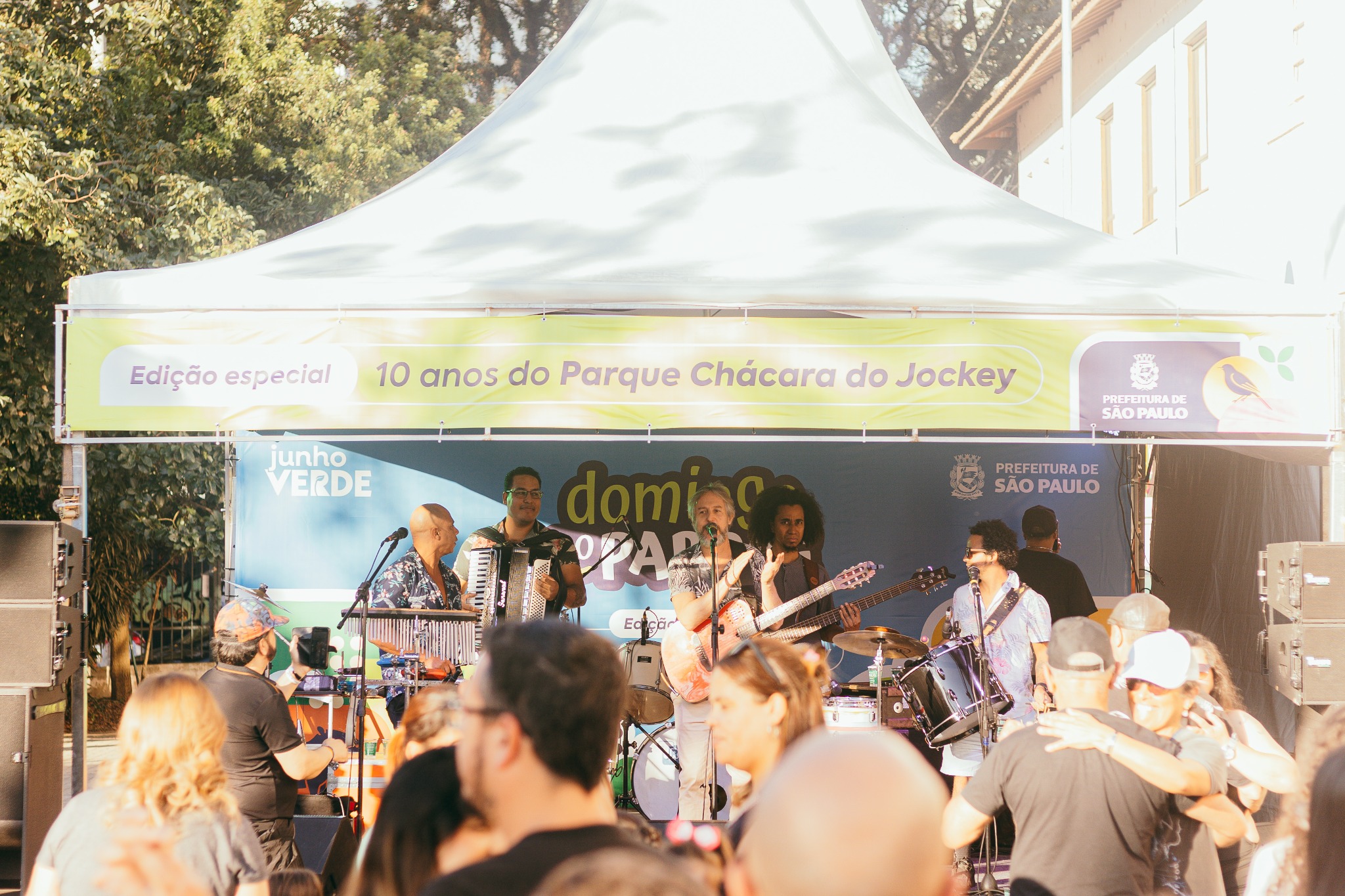 Fotografia colorida registrada ao ar livre durante o dia, em um evento realizado no Parque Chácara do Jockey. Ao centro da imagem, há um palco montado sob uma grande tenda branca. Na parte superior da estrutura, uma faixa em tons de amarelo e verde-claro traz a inscrição “Edição especial – 10 anos do Parque Chácara do Jockey”, acompanhada dos logotipos da Prefeitura de São Paulo. No palco, uma banda se apresenta para o público. Os músicos utilizam diferentes instrumentos, como violão, acordeão, baixo, bateria e percussão. Ao fundo, um painel azul exibe os dizeres “Domingo no Parque” e a identidade visual do Junho Verde.  Em primeiro plano, diversas pessoas acompanham a apresentação. Algumas estão em pé observando o show, enquanto outras conversam ou dançam. As roupas variam entre tons escuros, azuis, brancos e estampados coloridos. A iluminação do fim de tarde cria áreas de luz dourada sobre o público e o palco. Ao redor da tenda, destacam-se árvores de folhagem verde intensa, que ocupam grande parte do fundo da imagem, reforçando o ambiente de parque e lazer ao ar livre. À direita, é possível ver parte de um edifício de fachada clara, em tons de branco e bege.