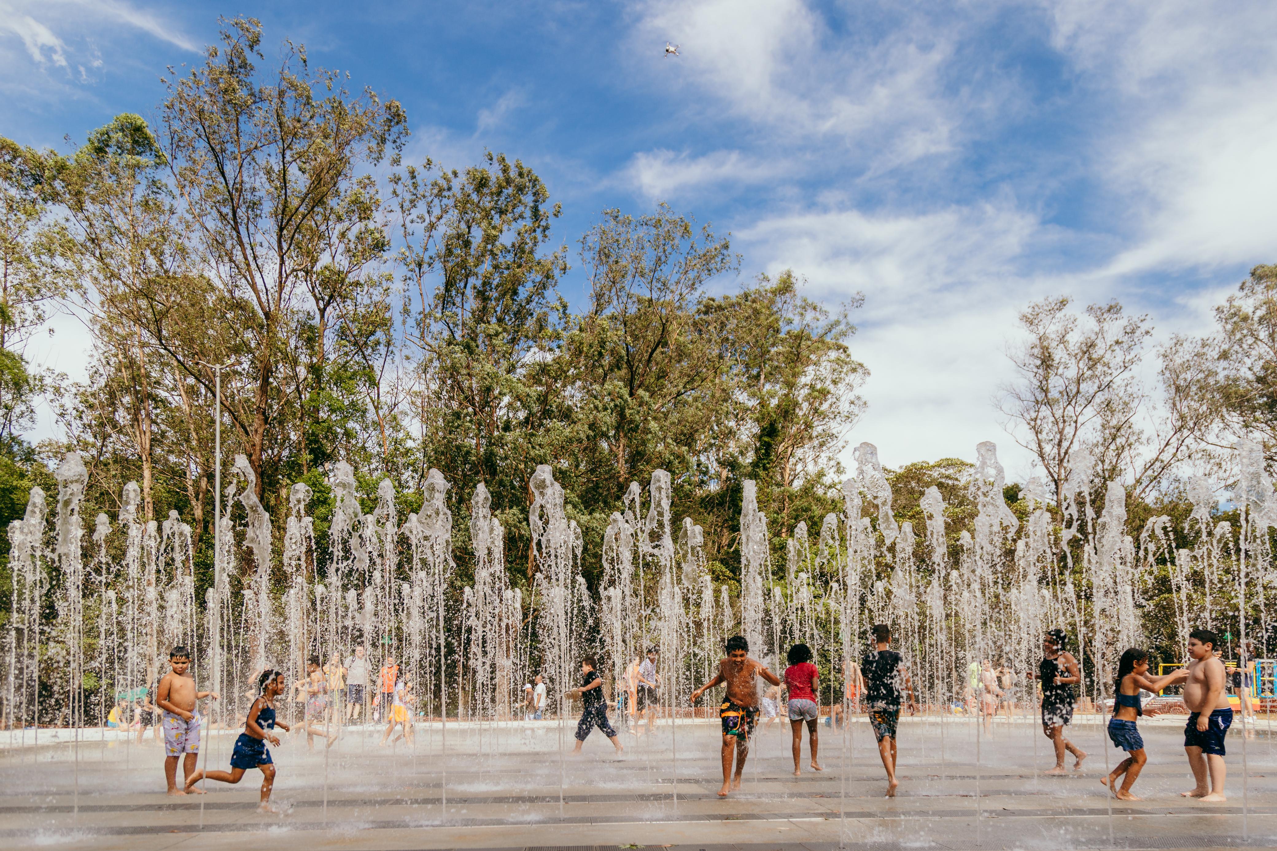 A imagem mostra uma grande fonte interativa ao ar livre, com dezenas de jatos de água transparentes subindo em diferentes alturas. As gotas brilham sob a luz do sol, criando reflexos prateados e brancos. À frente da fonte, várias crianças correm e brincam molhadas; elas usam roupas de banho coloridas, como azul, rosa, laranja e roxo.  Ao fundo, há uma área verde composta por árvores altas de troncos marrons e folhagens em tons de verde claro e verde escuro. Algumas pessoas observam a brincadeira ao longe, também vestindo roupas variadas e coloridas. O céu ocupa a metade superior da imagem, com um azul intenso mesclado a nuvens brancas suaves e esfiapadas, dando um aspecto luminoso e agradável à cena.