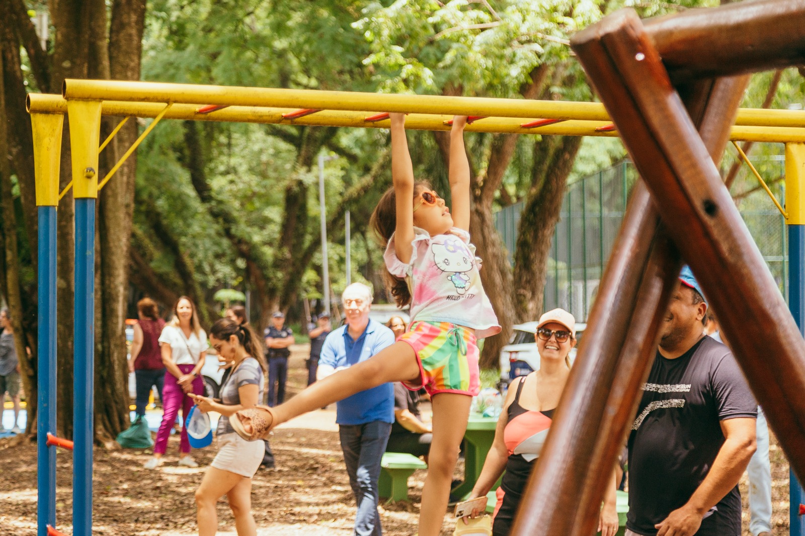 Criança brinca em brinquedo de barras em um parque, enquanto adultos observam ao fundo.