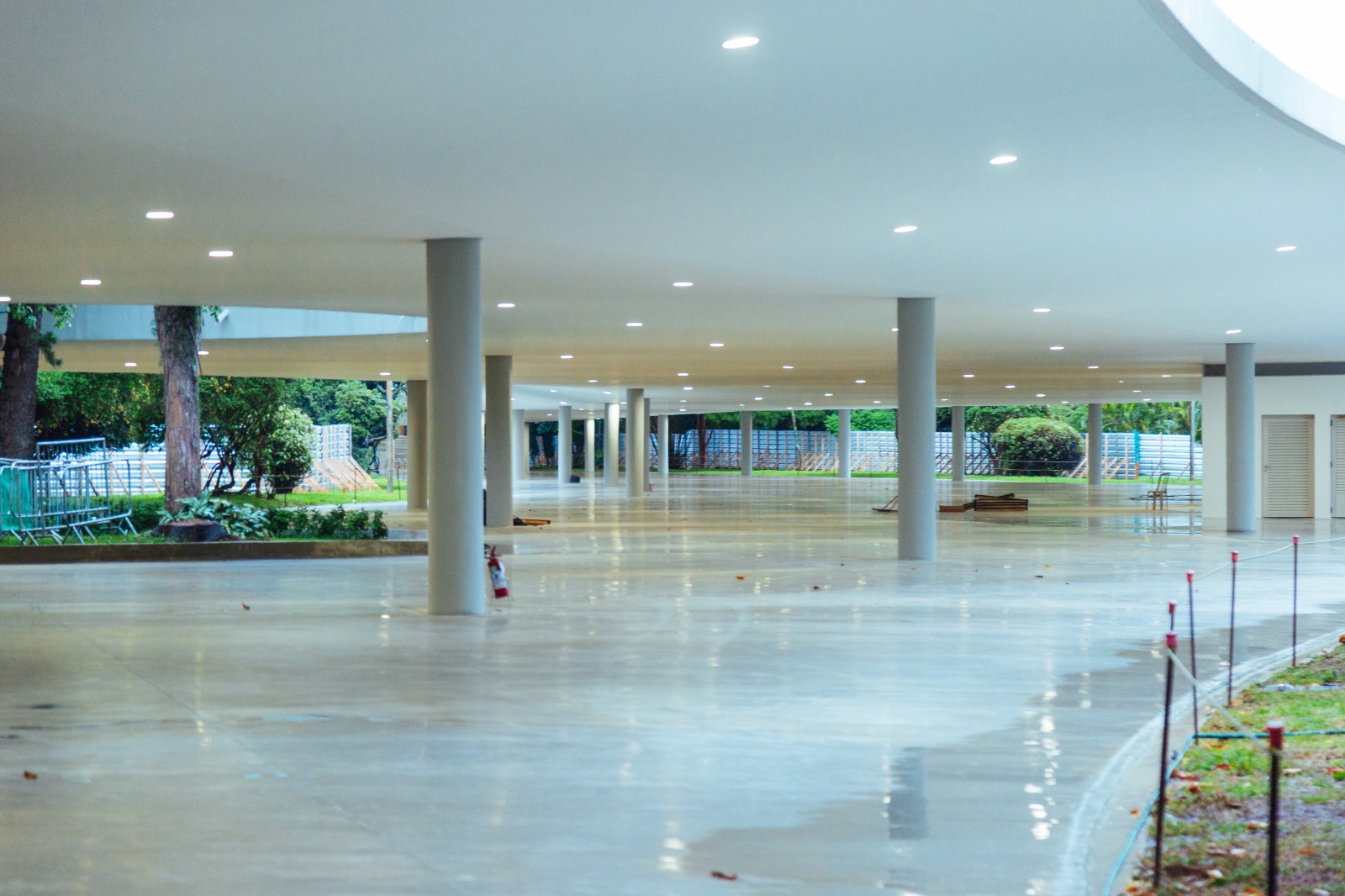 Imagem do interior da Marquise do Ibirapuera. O espaço é amplo, coberto e aberto nas laterais, com piso liso e claro que reflete a iluminação do teto. O teto branco é baixo e contínuo, com várias luminárias circulares embutidas, distribuídas de forma regular. Diversas colunas cilíndricas cinza sustentam a estrutura, alinhadas em fileiras que criam profundidade. À esquerda e ao fundo, é possível ver áreas verdes do parque, com árvores, arbustos e grades de proteção temporárias. À direita, há uma faixa de jardim acompanhando o contorno do piso, delimitada por pequenas estacas com corda. O espaço está vazio, transmitindo sensação de amplitude, organização e arquitetura moderna integrada ao parque.