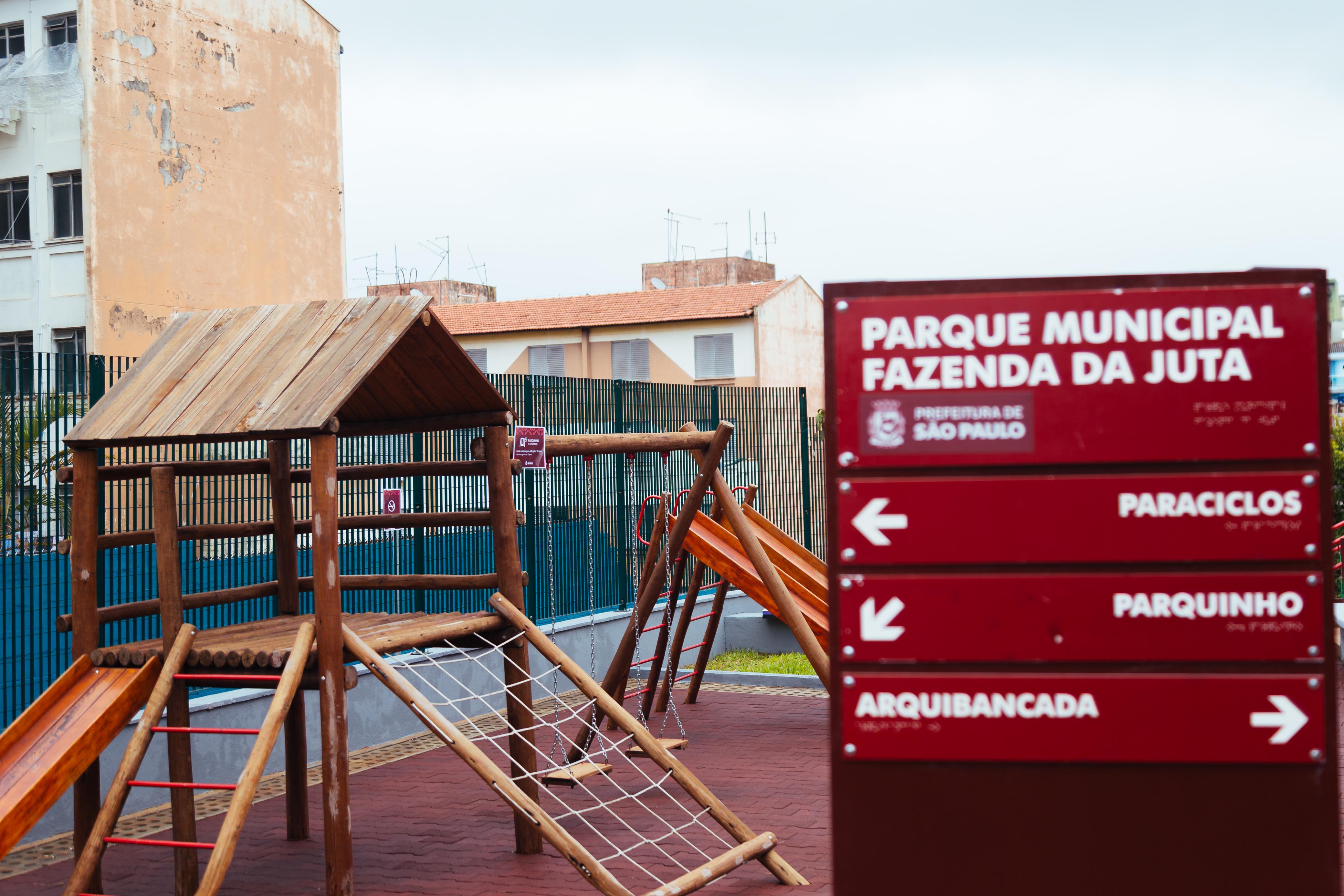 Imagem diurna de um parquinho infantil em um espaço urbano. À esquerda, há um brinquedo de madeira com cobertura, escorregador, escada e uma rede de cordas para escalar. O piso é emborrachado na cor avermelhada. Ao fundo, vê-se uma grade verde e prédios residenciais. À direita, em primeiro plano, há uma placa vermelha com a inscrição “Parque Municipal Fazenda da Juta – Prefeitura de São Paulo”, com setas indicando os espaços: Paraciclos, Parquinho e Arquibancada.