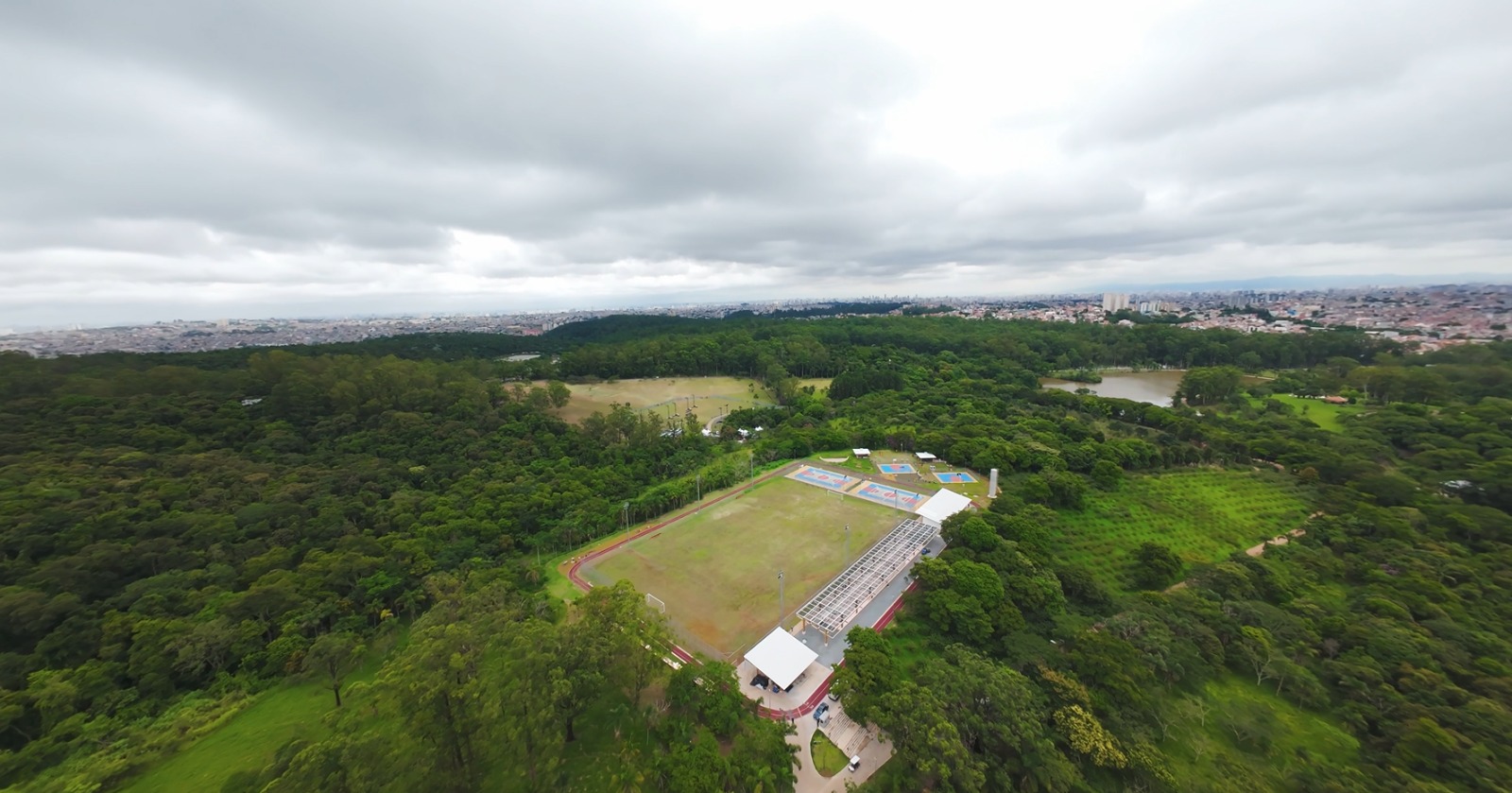 Imagem aérea de um grande parque urbano cercado por extensa área verde. No centro, há um complexo esportivo com um campo de futebol gramado, pista de atletismo ao redor e arquibancada coberta com estrutura metálica clara. Ao lado do campo, aparecem quadras poliesportivas coloridas em tons de azul e laranja. O parque é composto por muitas árvores densas, formando uma grande mancha verde. Ao fundo, vê-se a cidade, com inúmeros prédios e casas, sob um céu nublado e cinza.