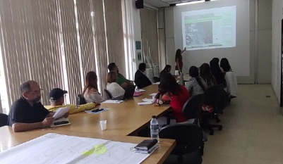 Sala de reunião iluminada, com uma mesa longa de madeira ao centro. Cerca de dez pessoas estão sentadas ao redor, algumas com papéis e anotações à frente. No fundo da sala, uma mulher em pé apresenta um conteúdo projetado na parede, que mostra um slide com imagens e textos. À esquerda, há janelas grandes com persianas parcialmente fechadas, deixando entrar luz natural. O ambiente é organizado e corporativo, sugerindo uma reunião ou treinamento.