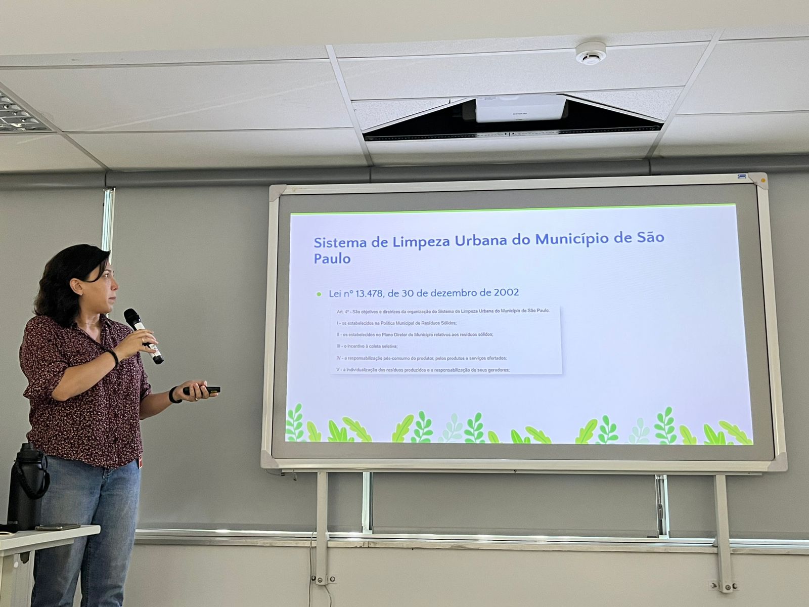 Mulher em pé apresenta um slide projetado em uma tela, segurando microfone e controle. O slide fala sobre o Sistema de Limpeza Urbana de São Paulo. Ambiente interno, tipo sala de aula ou reunião.