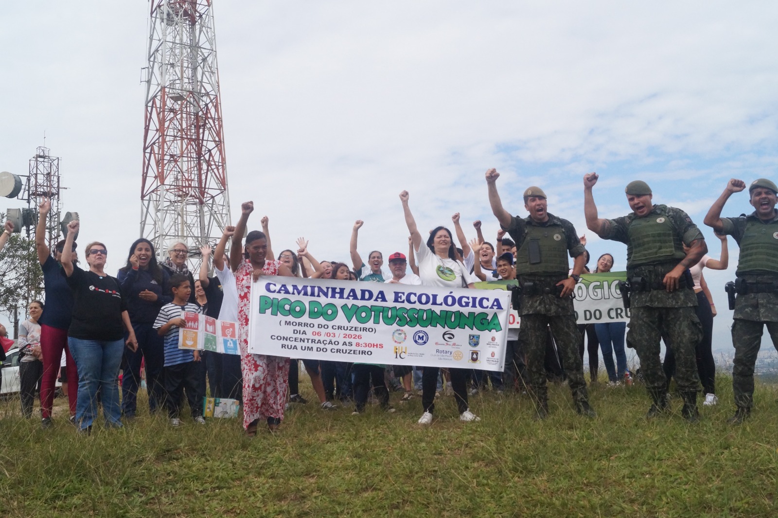 Grupo de pessoas participa de caminhada ecológica no Pico do Votussununga, em área de gramado no Morro do Cruzeiro. Os participantes aparecem reunidos segurando uma faixa do evento, enquanto levantam os braços em clima de celebração. Ao lado do grupo, agentes fardados também participam da atividade. Ao fundo, aparecem torres de comunicação e céu nublado.