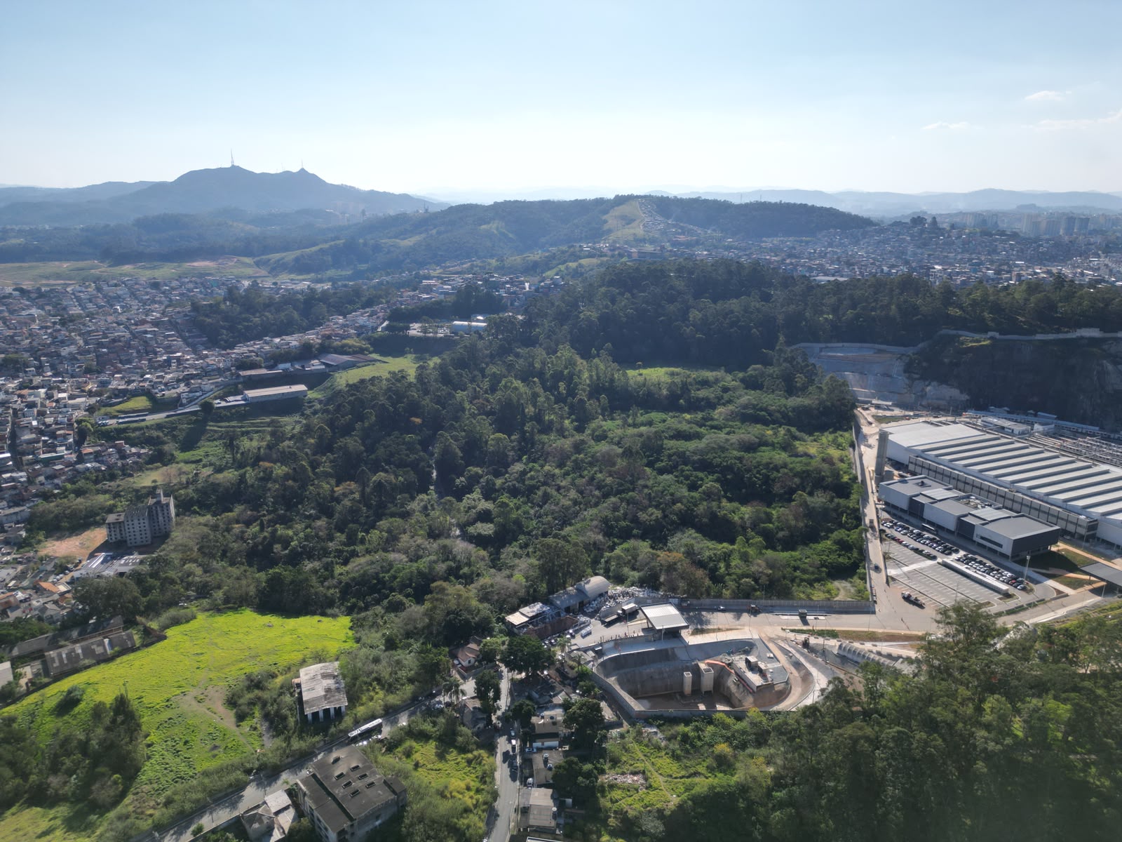 Vista aérea de uma área verde extensa, cercada por bairros, morros e construções urbanas. Há uma grande área de vegetação no centro e, à direita, um complexo de prédios industriais.
