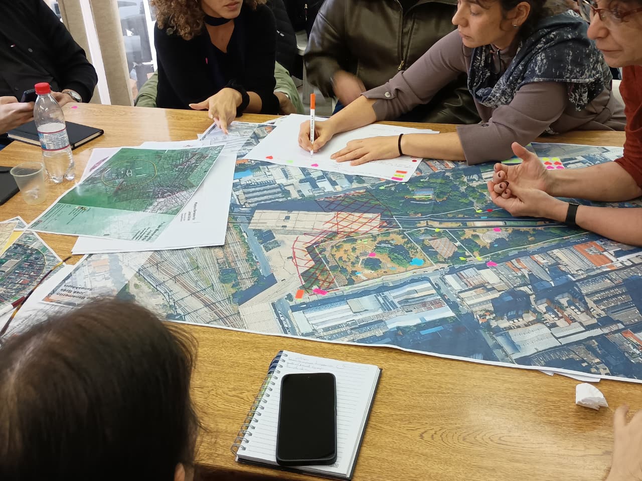 Participantes analisam mapas impressos sobre uma mesa e marcam diferentes áreas com canetas coloridas durante atividade colaborativa do SampaAdapta.