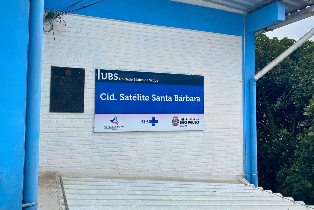 PraCegoVer: Fechada na UBS Cidade Satélite Santa Bárbara, com uma placa com os dizeres "Unidade Básica de Saúde Cid. Satélite Santa Bárbara" nas cores azul, preto e branco. As parede da UBS tem tijolinhos na cor branca com paredes na cor azul claro.