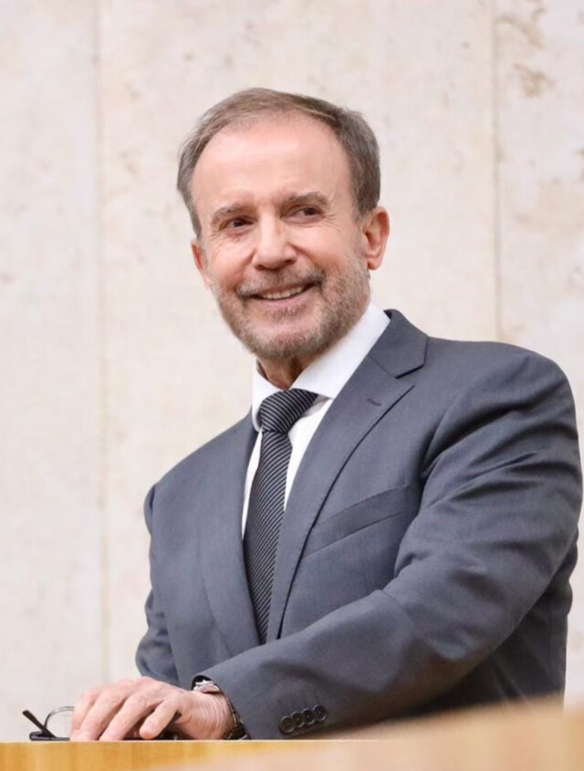 A imagem mostra um homem de meia-idade ou idoso, de cabelos curtos grisalhos e barba bem aparada, vestindo terno cinza escuro, camisa branca e gravata cinza com listras discretas. Ele está sorrindo levemente, com uma expressão cordial.  O homem parece estar apoiado em um púlpito ou bancada de madeira, com uma das mãos sobre a superfície. Ao lado da mão, há um par de óculos. A postura sugere que ele pode estar falando em público ou participando de um evento formal.  O fundo é composto por uma parede clara de pedra ou mármore, que transmite um ambiente institucional ou solene, possivelmente um edifício público ou local de cerimônia. A iluminação é suave e natural, destacando o rosto e o traje do homem.