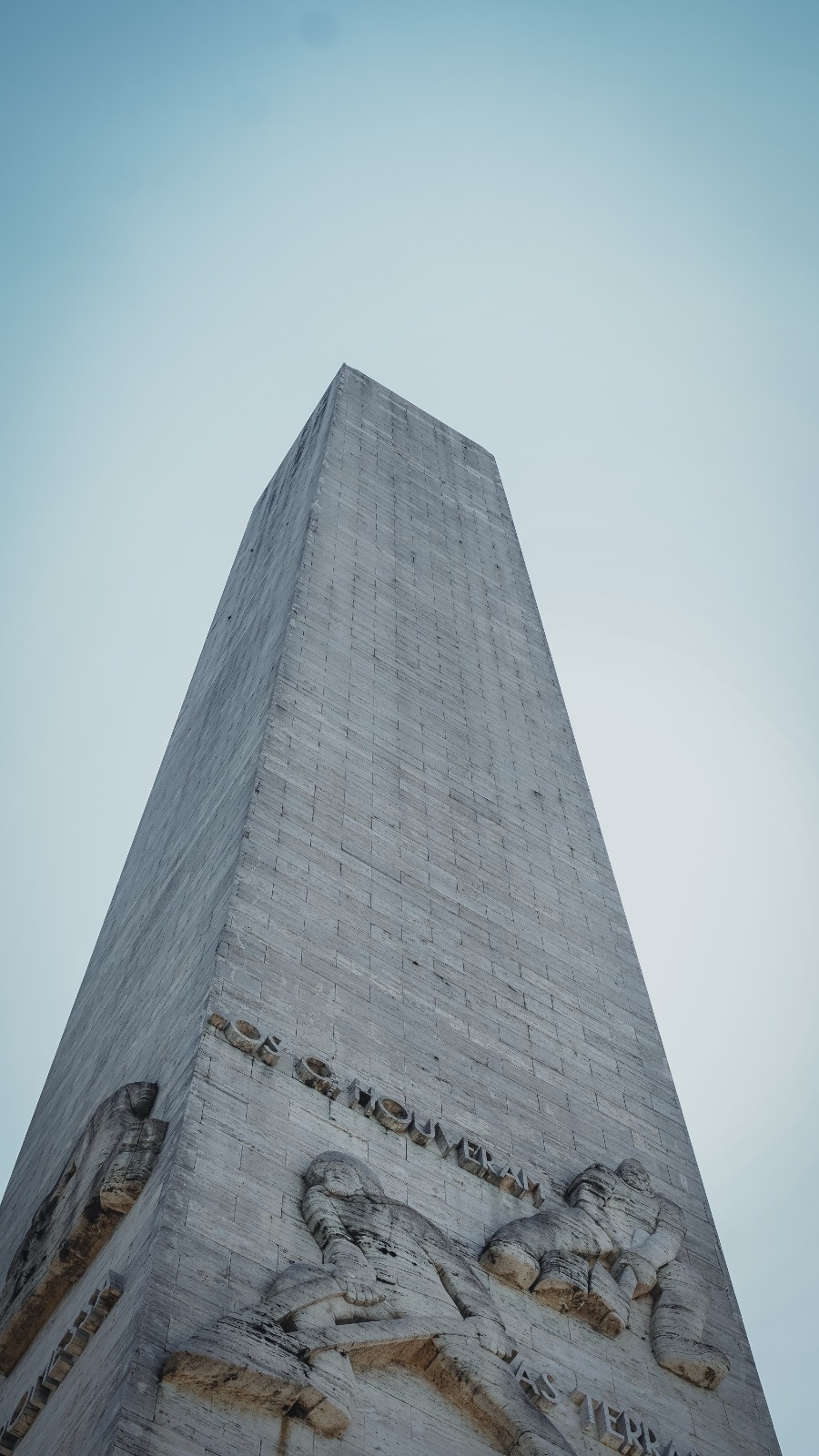 Monumento alto em pedra clara, visto de baixo para cima, com formato retangular e linhas retas, destacando esculturas em relevo de figuras humanas na parte inferior da estrutura; o céu aparece claro ao fundo, sem nuvens, reforçando a imponência do monumento.